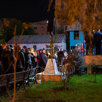 The image depicts a large group of people gathered in an outdoor area, possibly at night. They are standing around and socializing with each other. There is a bench located near the center of the scene, providing seating for some of the individuals present.