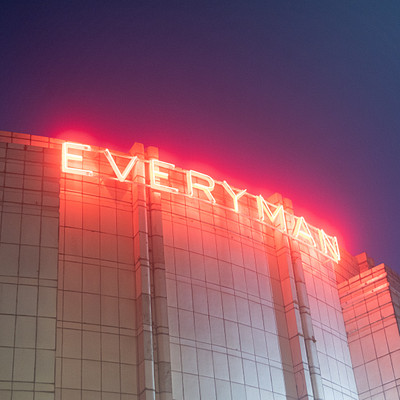 The image features a large building with a red neon sign that reads "EVERYMAN." The sign is illuminated, making it stand out against the dark sky. The building appears to be made of glass and stone, giving it an elegant appearance.