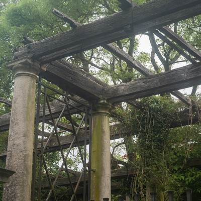 The image features a large stone structure with an open-air roof, possibly a gazebo or a pavilion. The roof is supported by wooden beams and has a vine growing on it. There are several trees surrounding the area, adding to the natural ambiance of the scene.