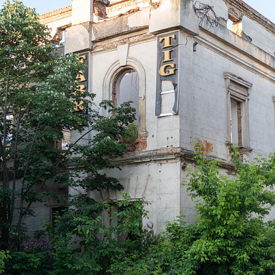 The image features a large, old building with a white facade. It appears to be an abandoned or neglected structure, as it is covered in ivy and surrounded by trees. A sign on the side of the building reads "TG." The building's overall appearance suggests that it has been left unused for quite some time.