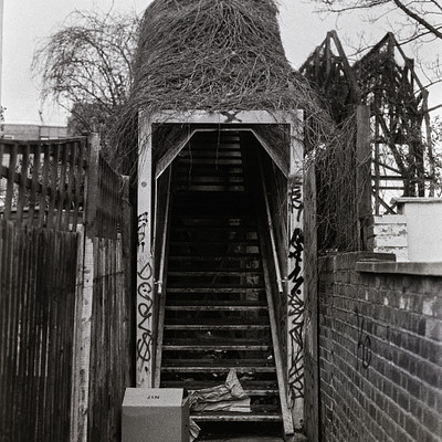 The image is a black and white photo of an old staircase leading up to a building. There are several steps visible in the scene, with some located closer to the bottom left side of the image and others nearer to the center. A box can be seen on the ground next to the stairs, possibly containing items or supplies for the building's occupants.