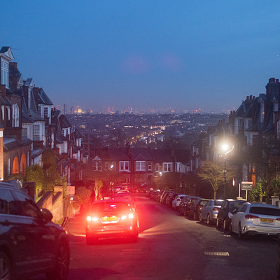 The image depicts a city street at night, with several cars driving down the road. There are multiple vehicles in various positions along the street, including some parked on the side of the road and others moving through the area. A few people can be seen walking or standing near the parked cars.