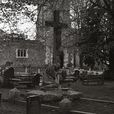 The image is a black and white photo of an old church with a large cross on top. The cross is located in the middle of the scene, surrounded by trees. There are several benches placed around the area, some near the cross and others further away.