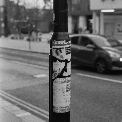 The image is a black and white photo of a street scene with a pole featuring an advertisement for a newspaper. A car is parked on the side of the road, and there are several people walking around in the area. Some individuals can be seen near the car, while others are scattered throughout the scene.