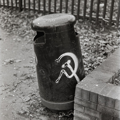 The image is a black and white photo of an old, dirty trash can with a hammer and sickle symbol on it. It appears to be sitting in the middle of a brick wall or fence. The trash can is located near a brick wall, which adds to the overall atmosphere of the scene.