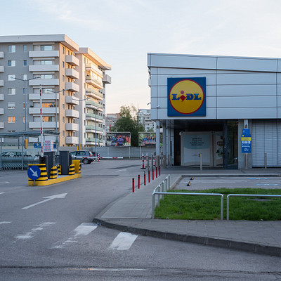 The image features a large blue building with the word "YEAH" on it, likely a supermarket. There are several cars parked outside of the building, and some traffic lights can be seen in the vicinity. A few people are walking around the area, possibly shopping or visiting the store.
