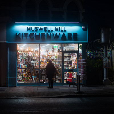 The image is a black and white photo of a kitchenware store at night. A man stands in front of the store, looking inside as he decides what to buy. There are several chairs placed around the store, with one near the left side, another on the right side, and two more towards the back.