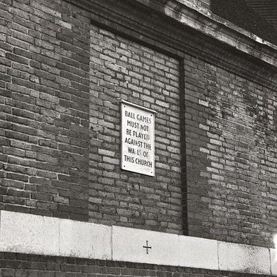 The image is a black and white photo of an old church with a sign posted on the side. The sign reads "ball games must be played against this of this church." The brick wall surrounding the church adds to its historical atmosphere.