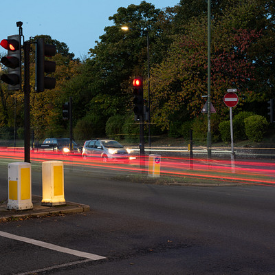 The image captures a busy street at night with multiple cars driving down the road. There are several traffic lights positioned along the street, some of which are red and others green. A few pedestrians can be seen walking on the sidewalk, adding to the bustling atmosphere.