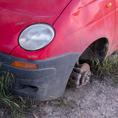 The image features a red car with its front tire off the ground, sitting in a grassy area. The tire is positioned on a rock and appears to be missing or damaged. The car's hood is open, revealing the engine underneath.