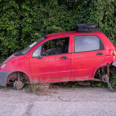 The image features a red car parked on the side of a road, with its hood up. It appears to be an old car that has been abandoned or is in need of repair. There are two tires visible underneath the car, one near the front and another towards the back.