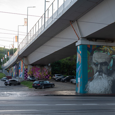 The image features a large, colorful mural painted on the side of a bridge. The mural is quite elaborate and captures attention with its vibrant colors and intricate design. In addition to the mural, there are several cars parked in the vicinity of the bridge.