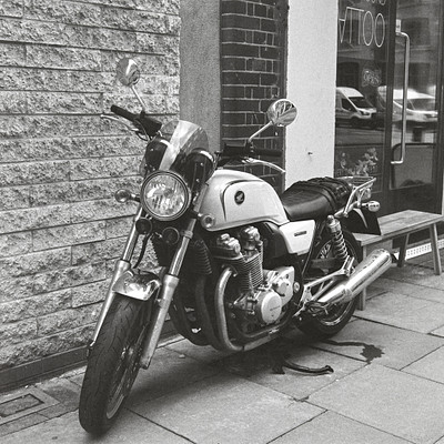 The image is a black and white photo of a motorcycle parked on the sidewalk. It appears to be an older model, possibly a vintage one. The motorcycle is positioned next to a brick wall, which adds a sense of depth to the scene.