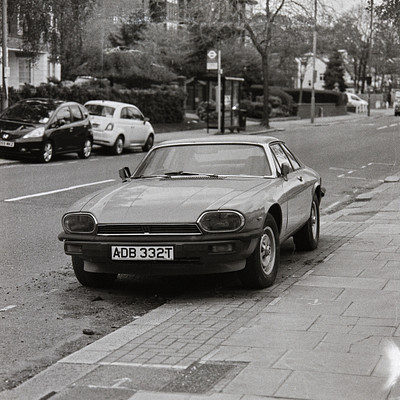 The image is a black and white photo of an old car parked on the side of a street. There are several other cars in the scene, including one behind the main car and two others further down the road. A bus can also be seen parked near the middle of the scene.