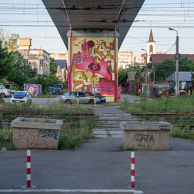 The image features a train crossing over a road, with a red bridge spanning the gap. A few cars are parked on both sides of the street, and there is a truck visible in the background. Additionally, there are several people walking around the area, some closer to the foreground while others are further away.