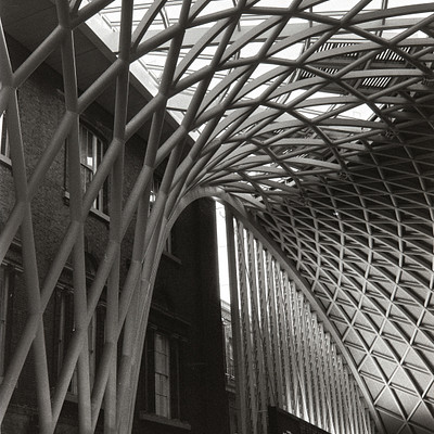 The image is a black and white photo of an old building with a large, curved metal structure above it. This structure appears to be a train station or a similar type of building. There are several arches in the scene, adding to the architectural interest.