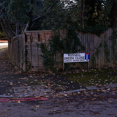 The image features a street sign on the side of a road, indicating that it is Rossell Close. There are trees in the background and a fence nearby. A small section of grass can be seen next to the fence.