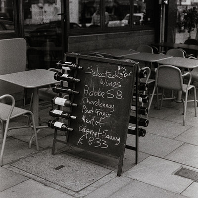 The image is a black and white photo of an outdoor restaurant with several tables and chairs. There are multiple wine bottles displayed on the sidewalk in front of the restaurant, possibly for sale or as part of a promotion. A chalkboard menu sign is also visible, indicating the types of wines available at this establishment.