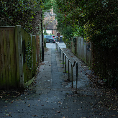 The image depicts a narrow alleyway between two houses, with a fence on either side. A car is parked at the end of the alley, and there are several benches placed along the path for people to sit and relax. Some of these benches are located near the fence, while others are closer to the middle of the alleyway.