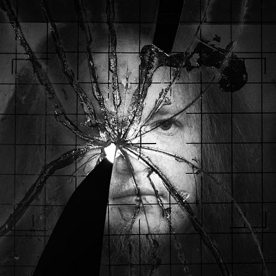 The image is a black and white photo of a man with a beard, looking directly into the camera. He appears to be staring through a hole in a piece of glass or a broken window pane. The man's face is prominently featured in the center of the picture, capturing his expression as he looks through the hole.