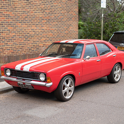 The image features a red and white striped sports car parked on the side of a road. It is positioned next to a brick wall, giving it an urban vibe. Another car can be seen in the background, further down the street.