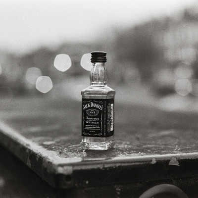 The image is a black and white photo of an open Jack Daniels bottle sitting on top of a trash can. The bottle appears to be empty, with the cap still in place. The scene takes place outdoors, possibly near a street or parking lot, as there are multiple cars visible in the background.