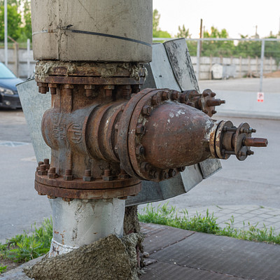 The image features a rusted fire hydrant sitting on the side of a road. It is located near a pole and appears to be in an old, run-down condition. There are several cars parked nearby, with one car positioned behind the fire hydrant and two others further away from it.
