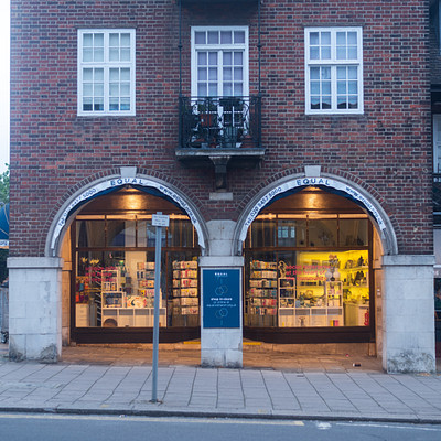 The image features a large brick building with two arched doorways, one on each side. Above the doors are signs that read "Books" and "Magazines." Inside the building, there is a bookstore filled with various books displayed in rows. In addition to the bookstore, there are several chairs placed throughout the scene, likely for customers to sit and relax while browsing through the selection of books.