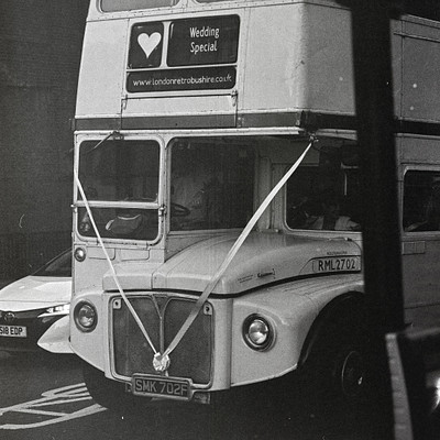 The image is a black and white photo of a double-decker bus with a wedding special sign on the front. The bus appears to be driving down a street, possibly in London. There are two people visible in the scene, one standing near the middle of the bus and another person closer to the right side.