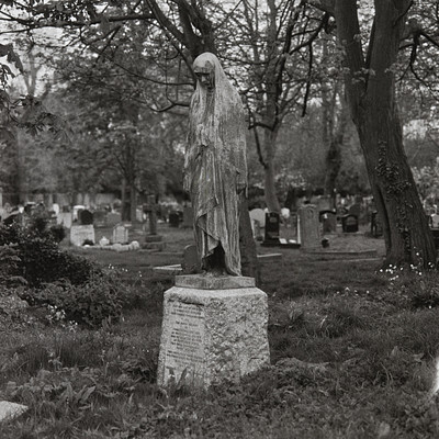 The image is a black and white photograph of a statue in a cemetery. The statue features a woman, possibly Mary, standing on top of a stone pedestal. She appears to be looking upwards or towards the sky.